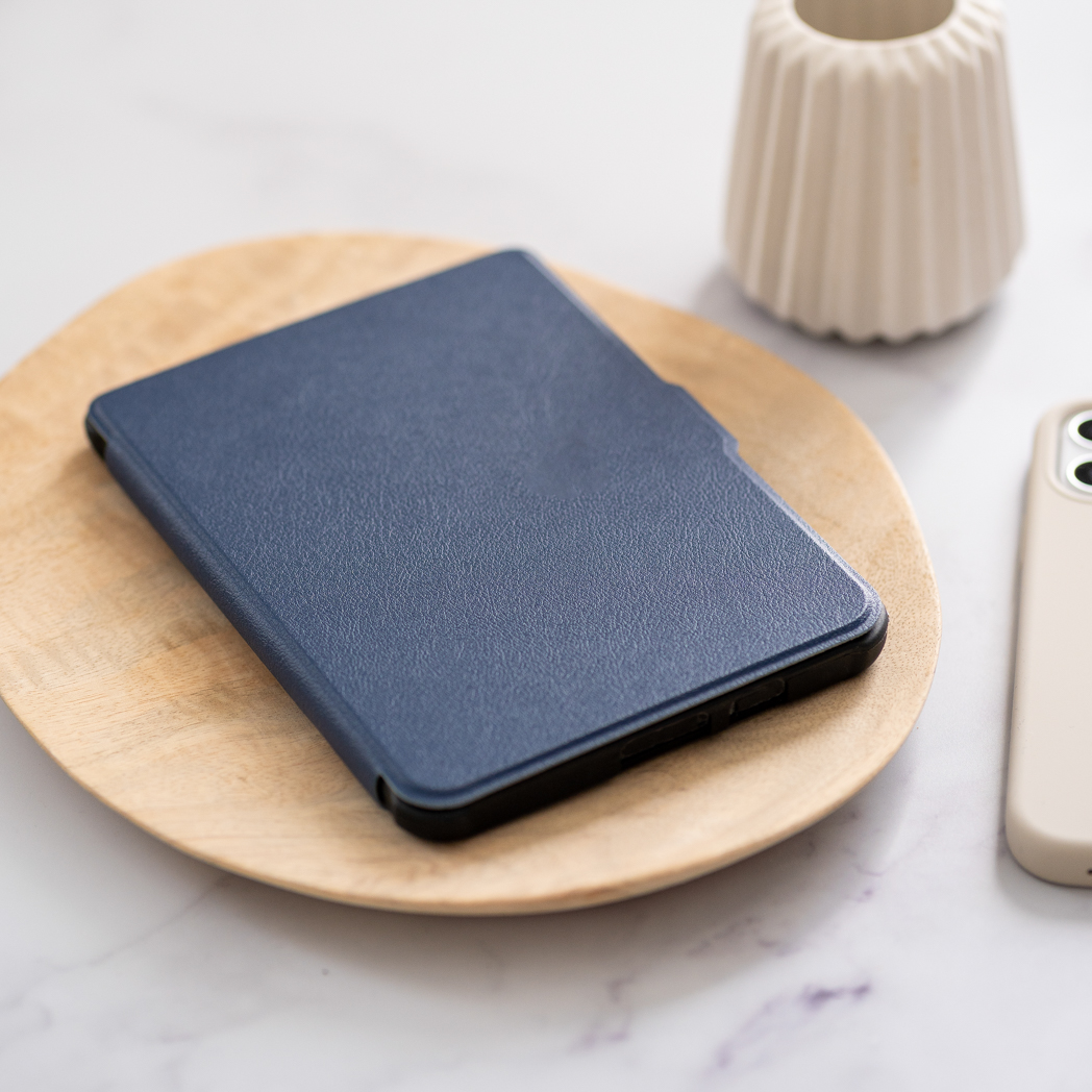 Un étui bleu foncé pour liseuse sur une assiette en bois. Un téléphone et un vase à proximité.