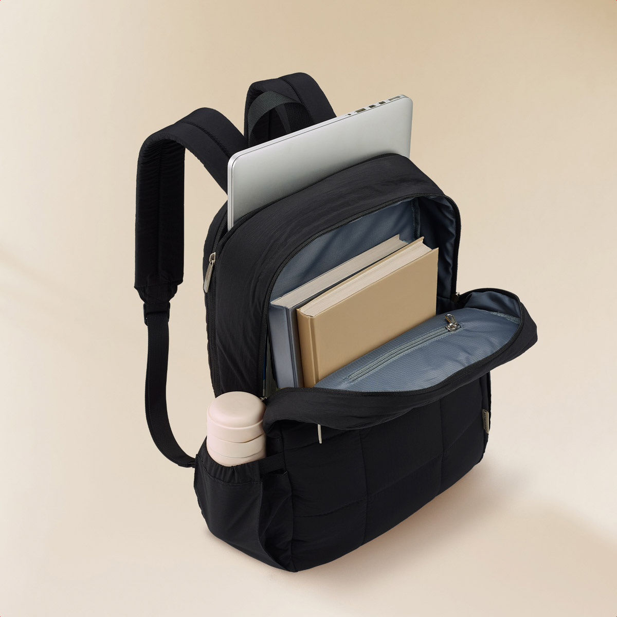 Black backpack with laptop, books, and cup. Open against a beige background.