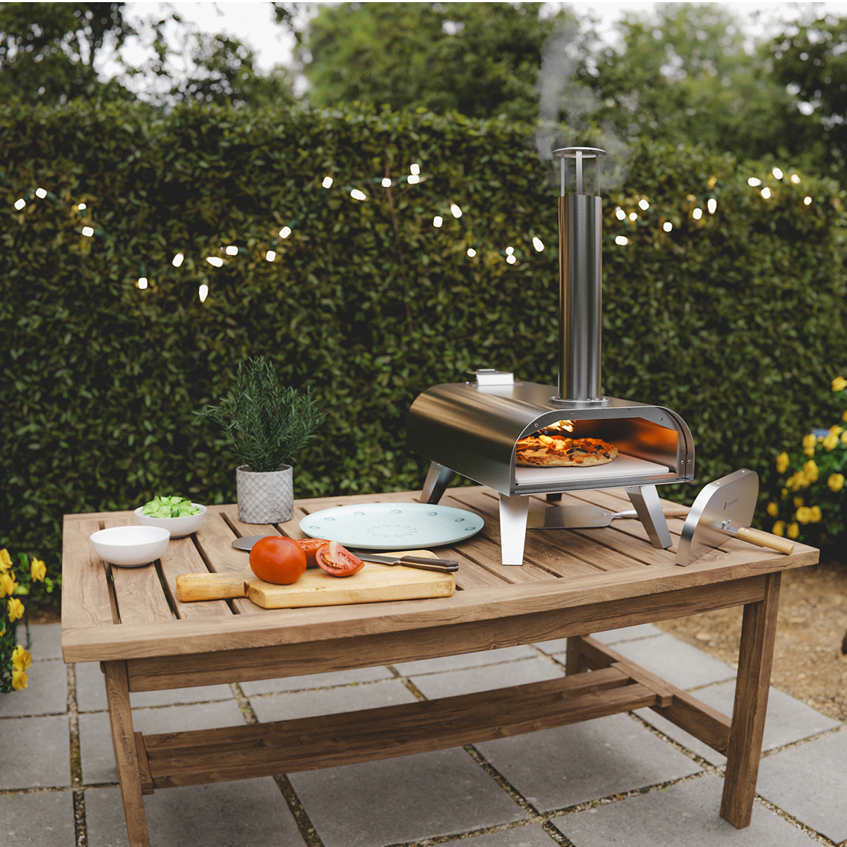 Een pizzaoven op een houten tafel met ingrediënten en lichtslingers op de achtergrond.