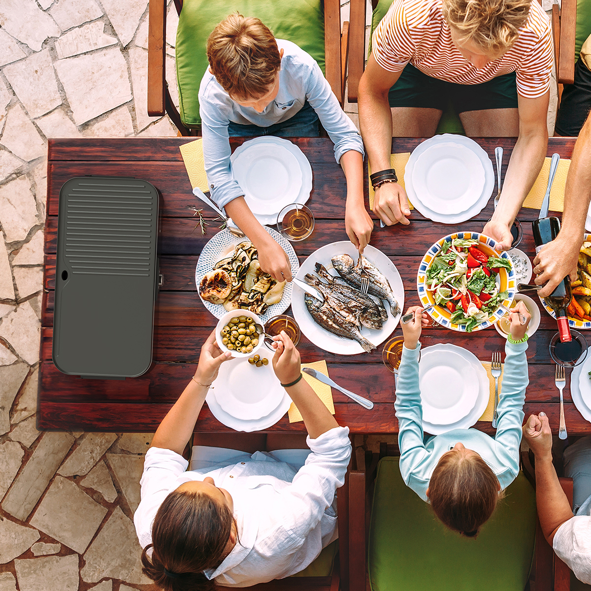 Family eating grilled fish, salad, and olives outdoors.