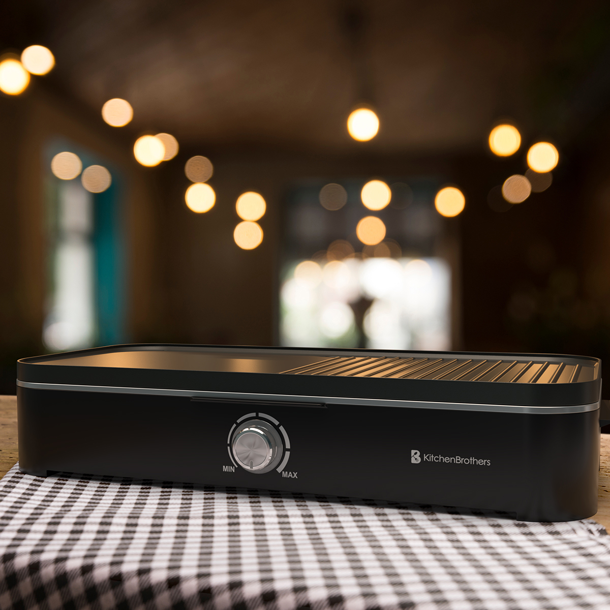 A black electric grill with a knob on a checkered cloth. Blurred lights in the background.