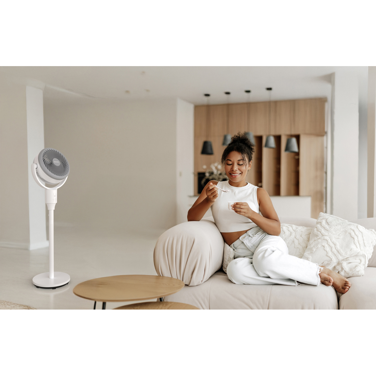 Eine Frau isst Joghurt auf einem Sofa, ein Ventilator steht im Hintergrund.