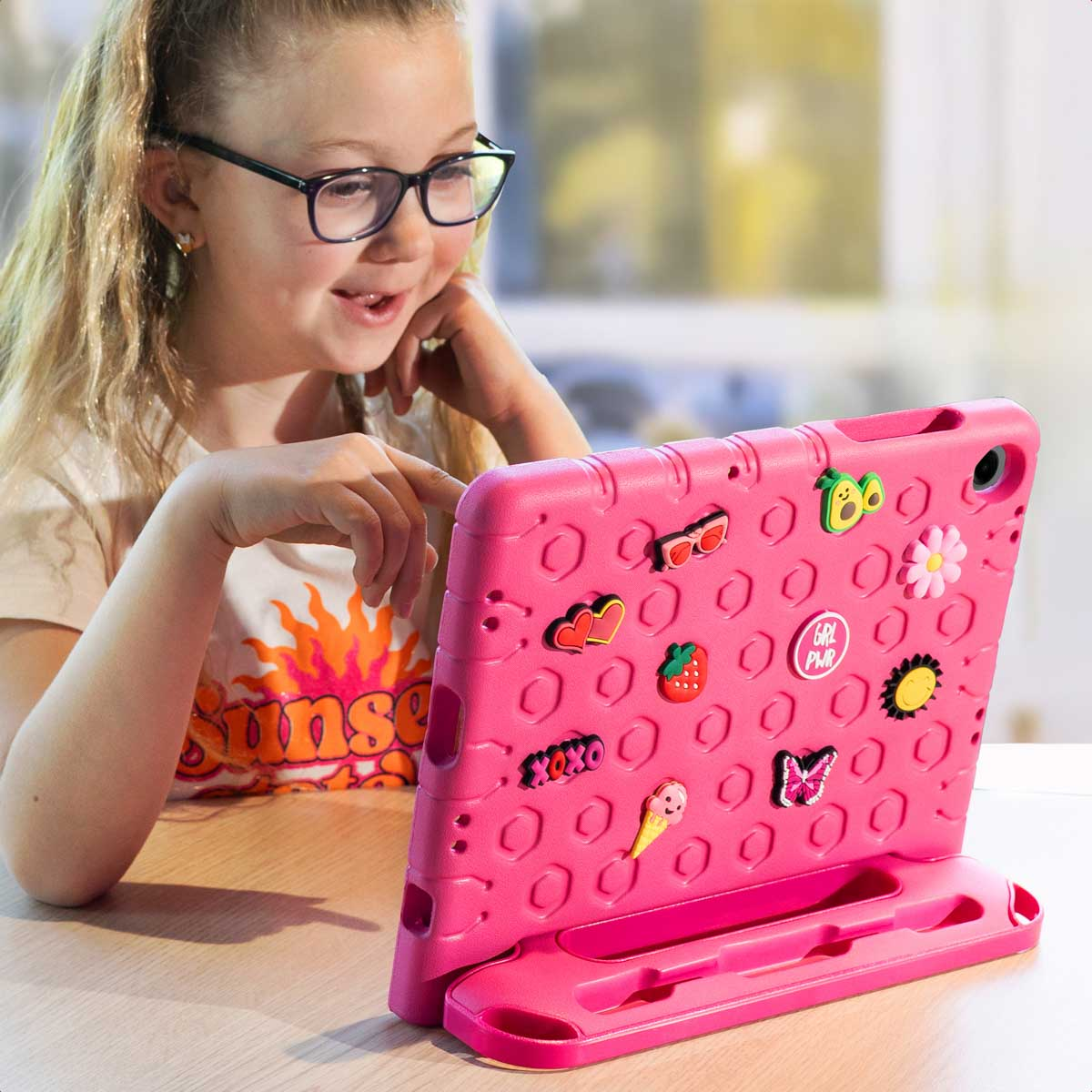 Een meisje met een bril kijkt lachend naar een roze tablet versierd met stickers.