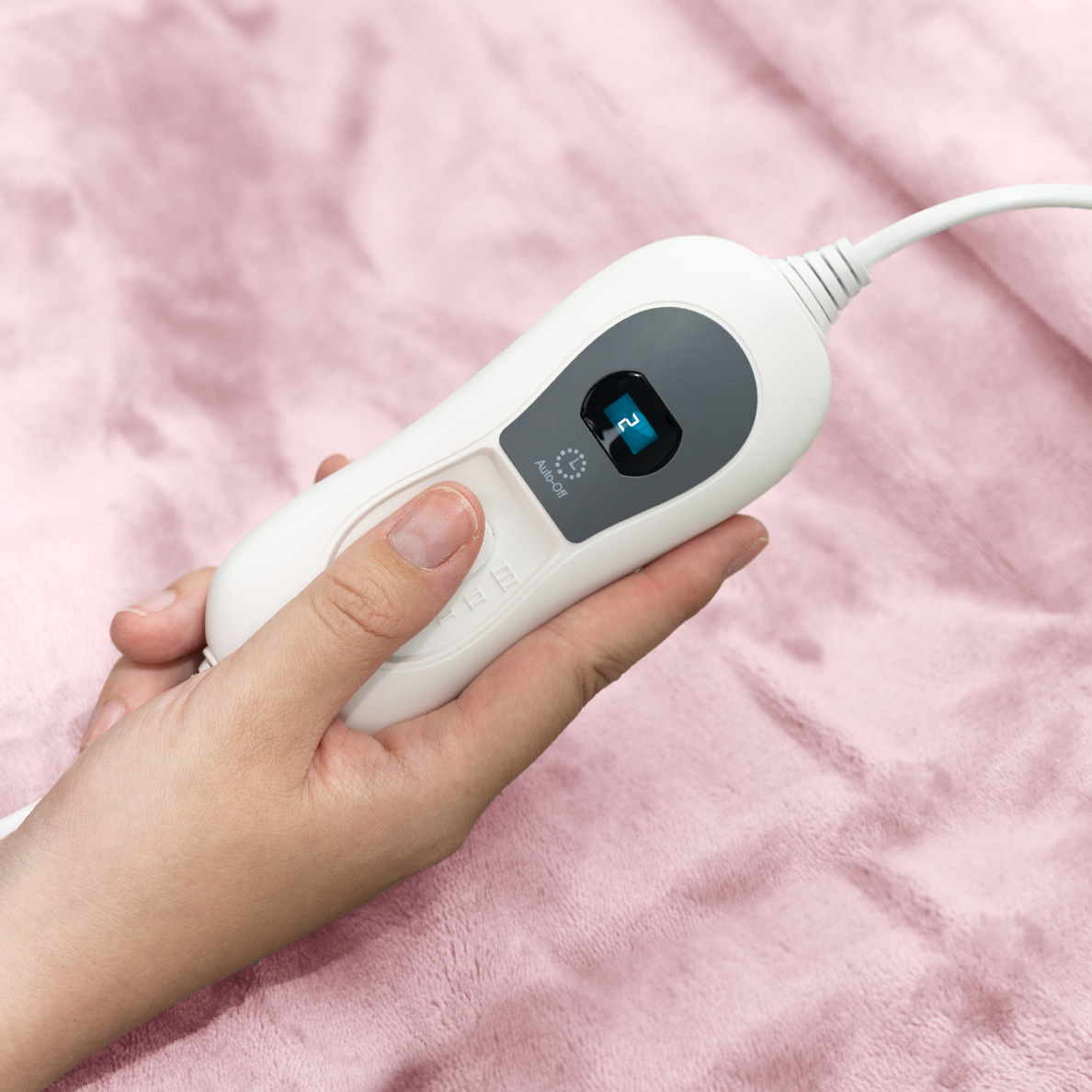 A hand holds a white electric blanket controller with a blue digital display on pink fabric.