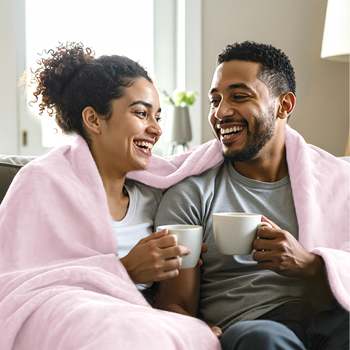 A couple shares a laugh while wrapped in a pink blanket, holding mugs.