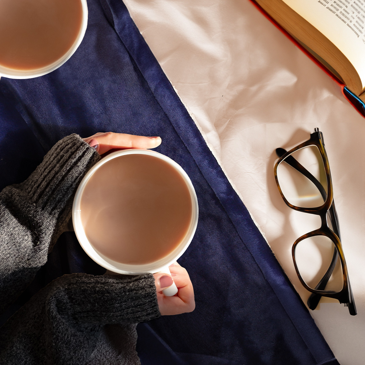 Hands hold a cup of cocoa next to a book and glasses.