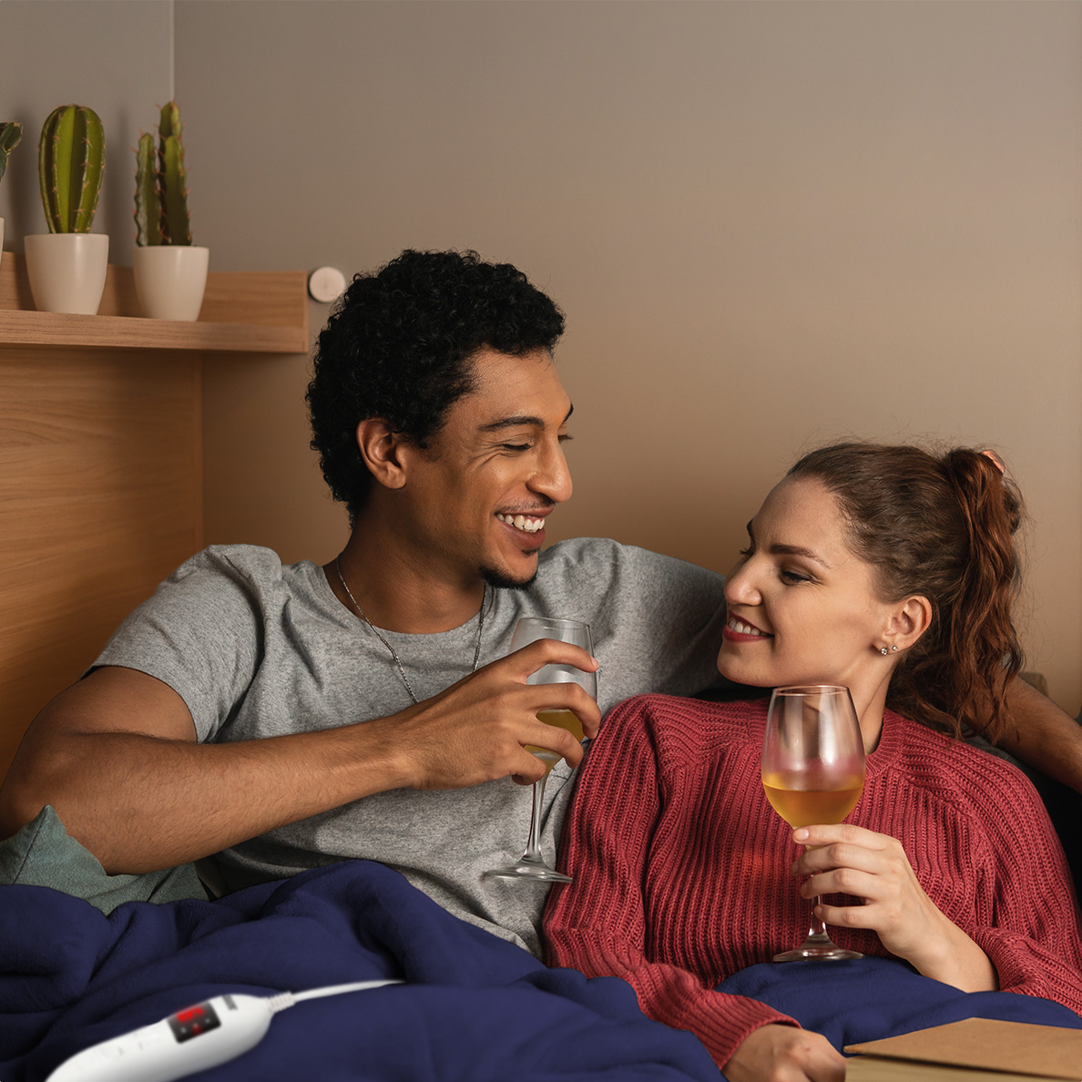 A man and a woman lie under a blanket, drinking wine.