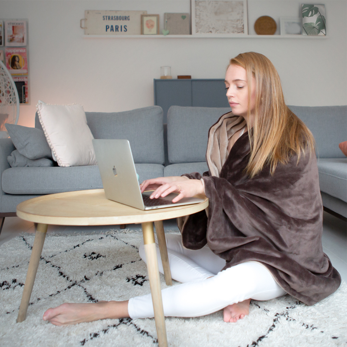 A woman in a brown cape sits on the floor using a laptop on a wooden table.