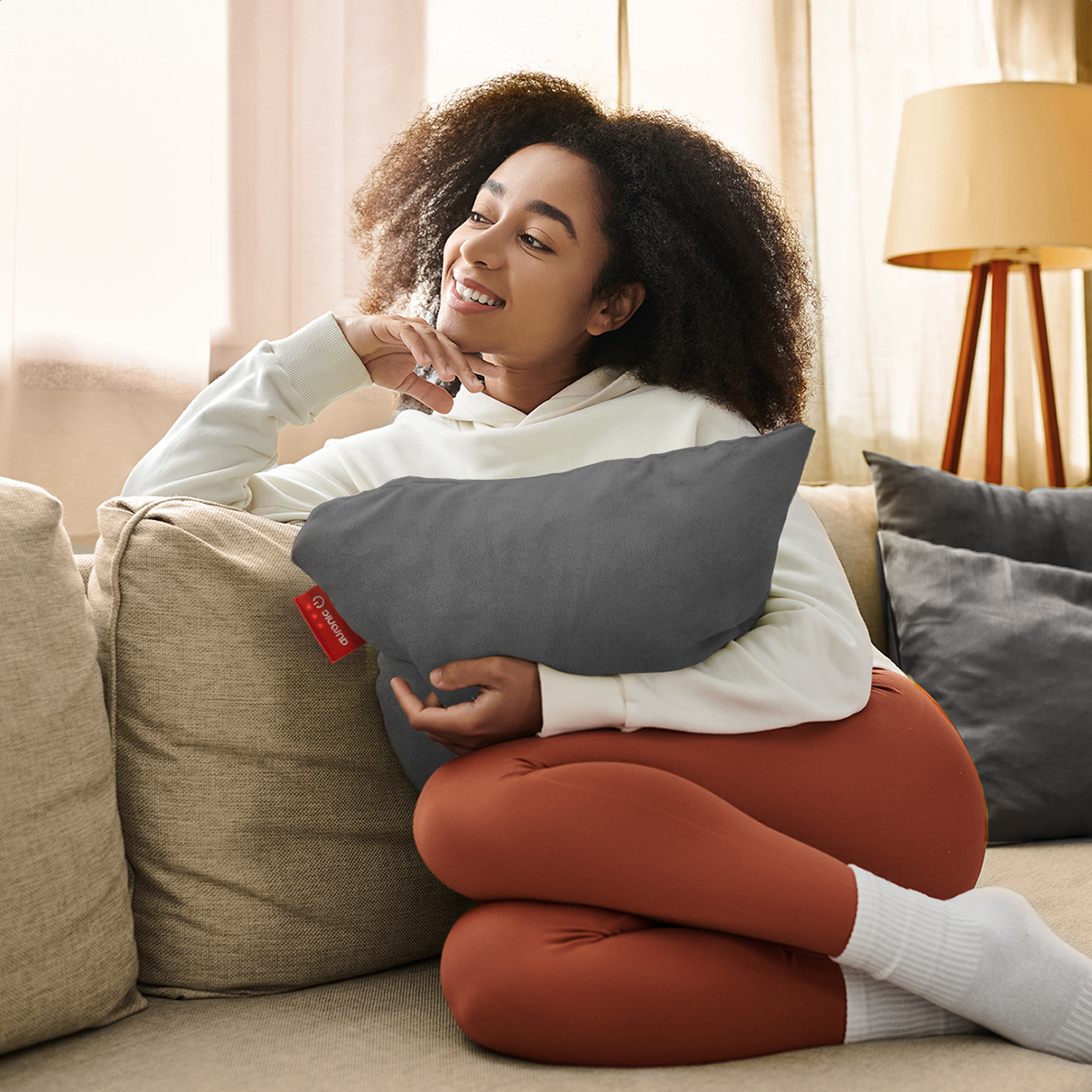 A woman sits on a sofa holding a gray pillow. She wears a white hoodie and orange leggings.
