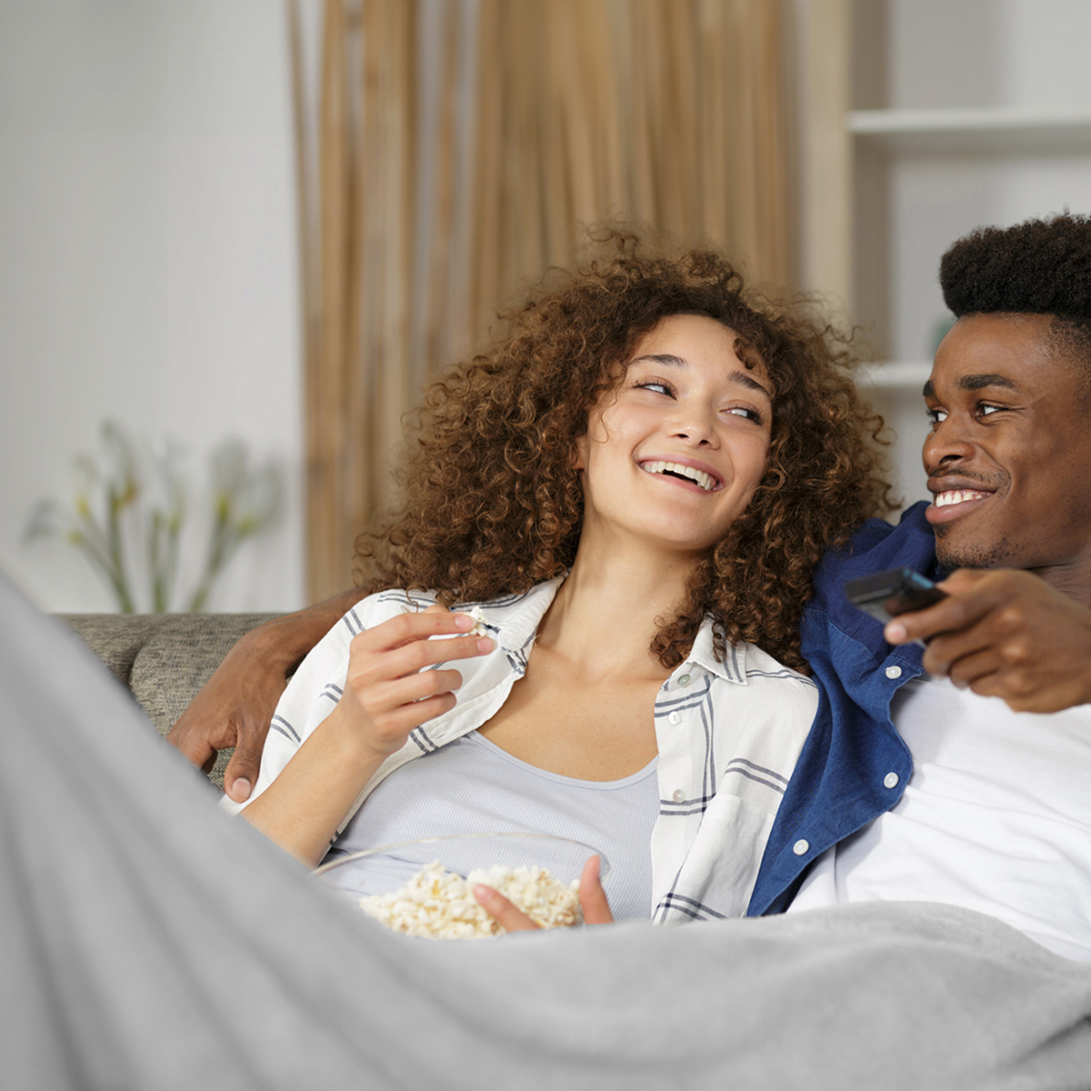 A couple on a couch eating popcorn and laughing while watching TV.