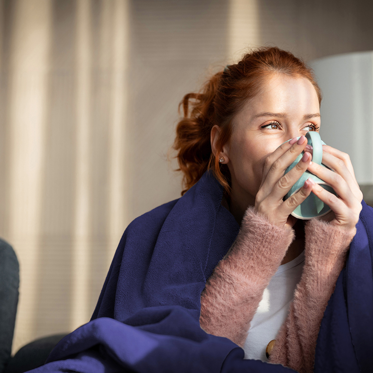 Une femme aux cheveux roux boit dans une tasse bleue, enveloppée dans une couverture.