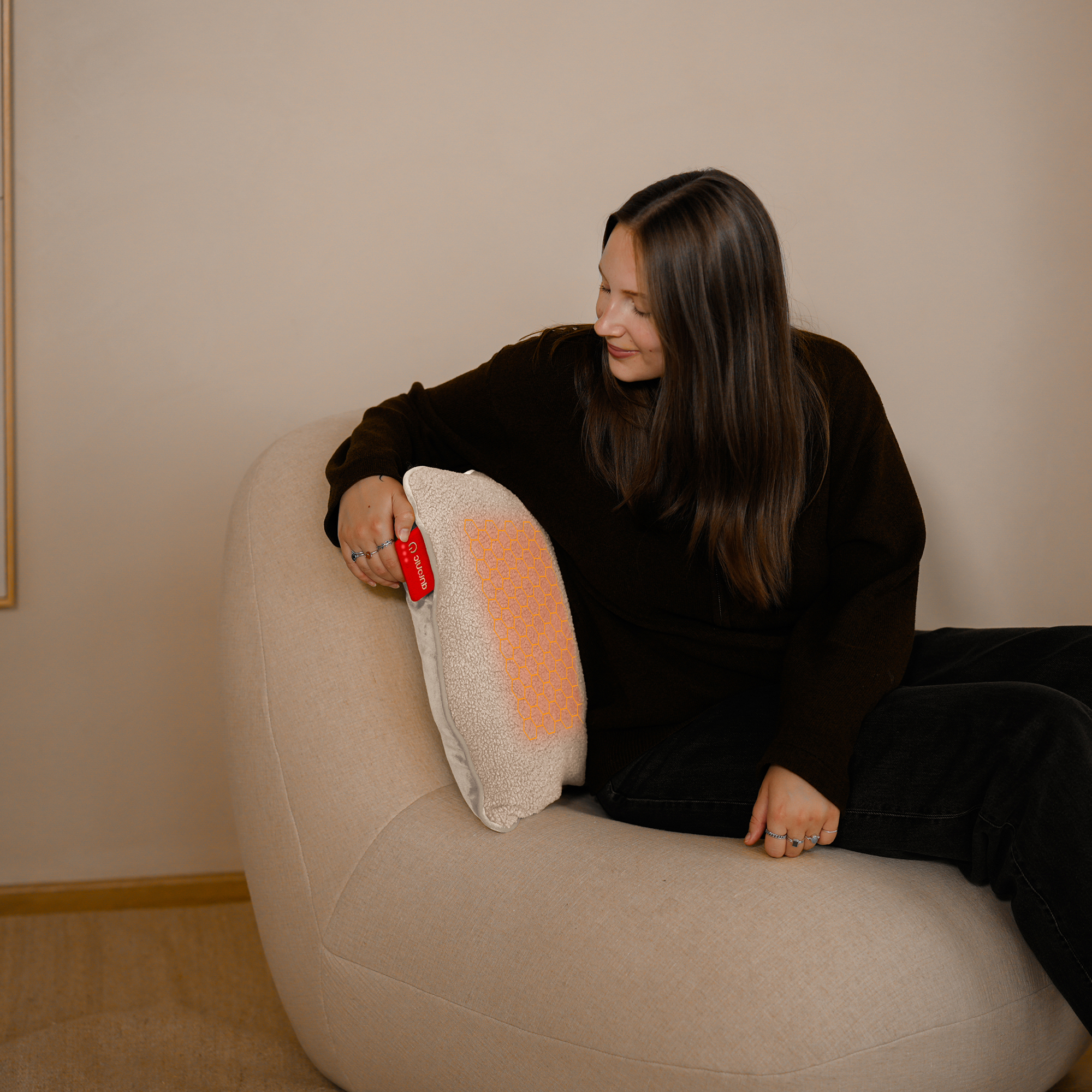 A woman sits on a beanbag chair holding a heated cushion with an orange pattern.