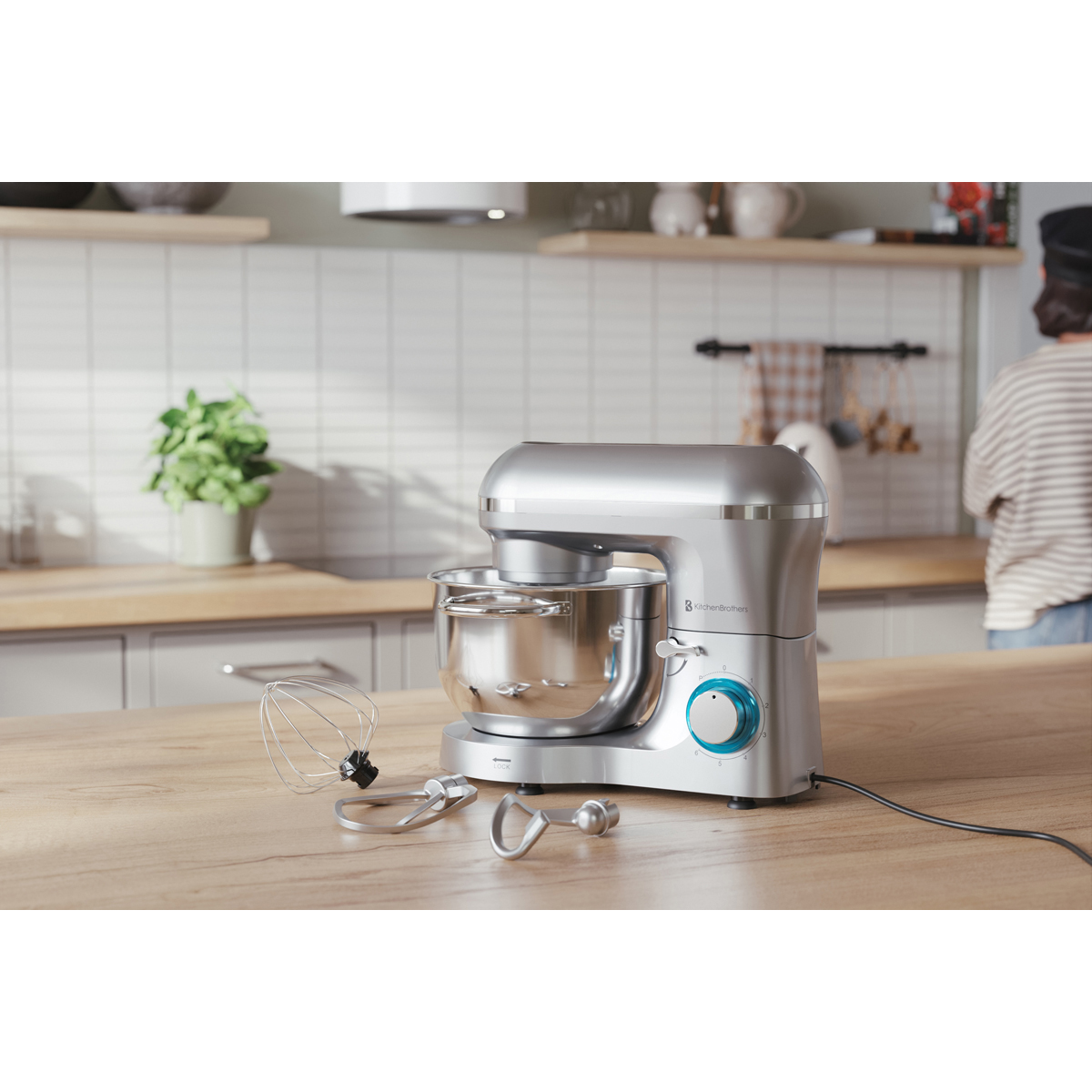 Een zilveren keukenmixer met kom en accessoires op een houten tafel.