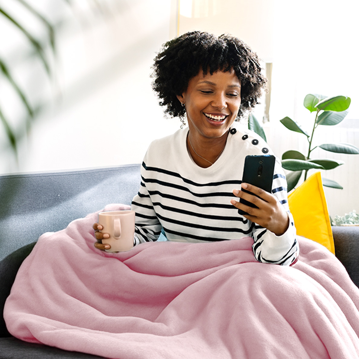 A smiling woman with curly hair sits on a couch holding a phone and a mug.
