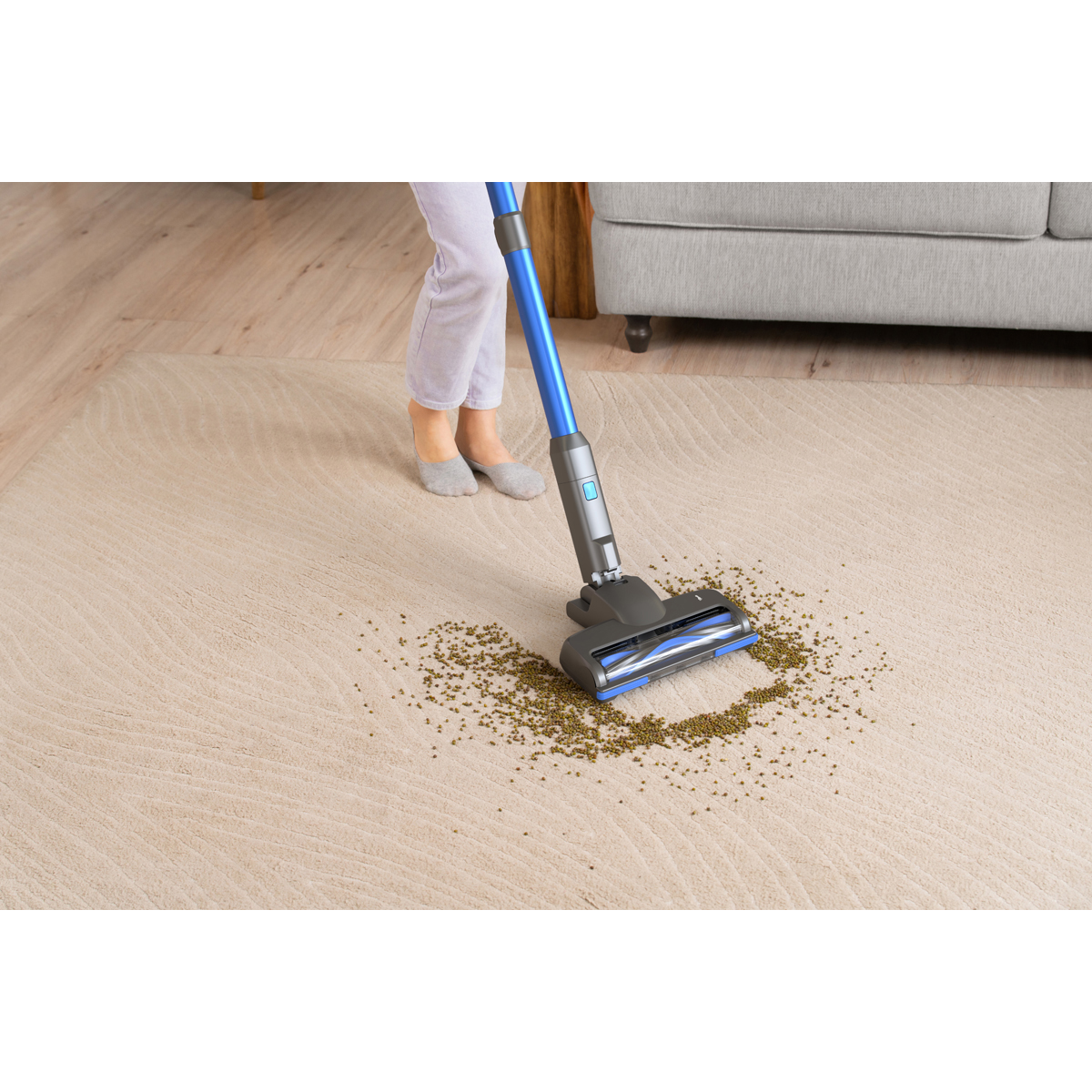 Une personne passe l'aspirateur sur des pois verts sur un tapis crème avec un aspirateur balai bleu.