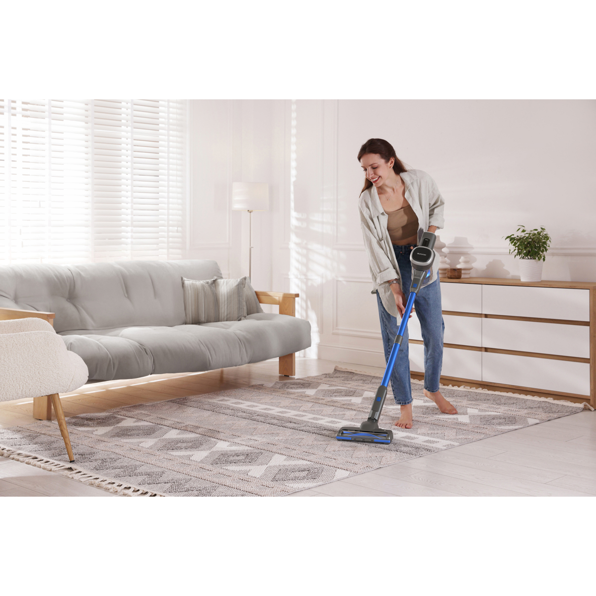 Une femme en jean et chemise passe l'aspirateur sur un tapis avec un aspirateur bleu.