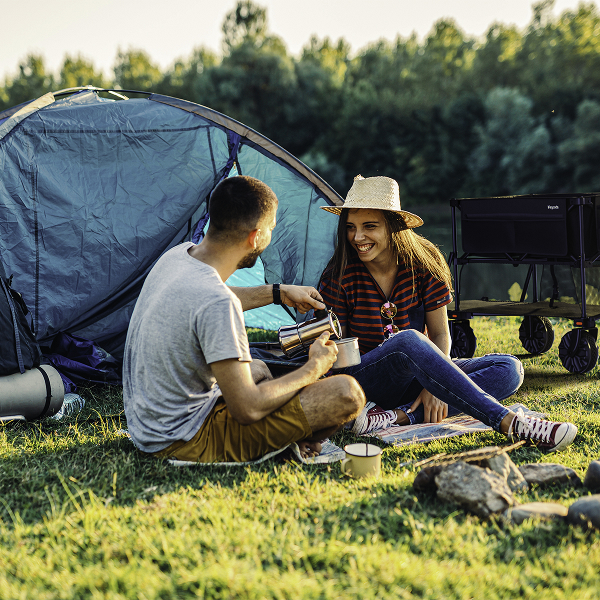 A man pours coffee for a woman at a campsite, with a cart in the background.
