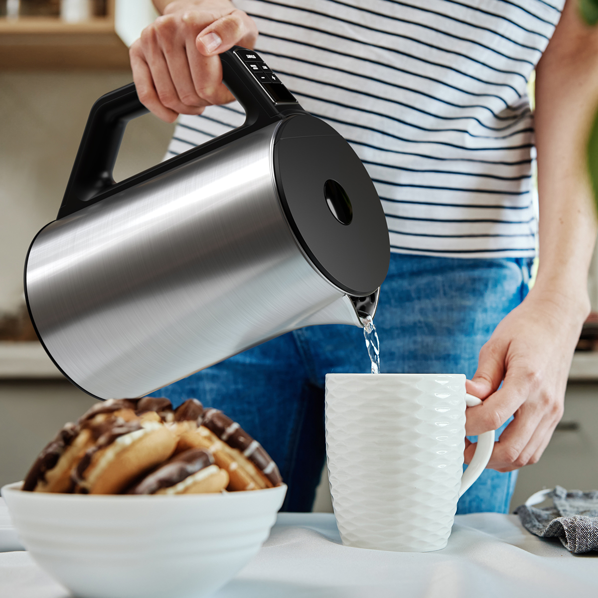 Personne versant de l'eau chaude d'une bouilloire dans une tasse, à côté d'un bol de biscuits.