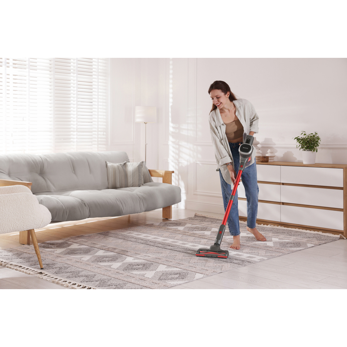 Une femme passe l'aspirateur sur un tapis à motifs dans un salon lumineux avec canapé.