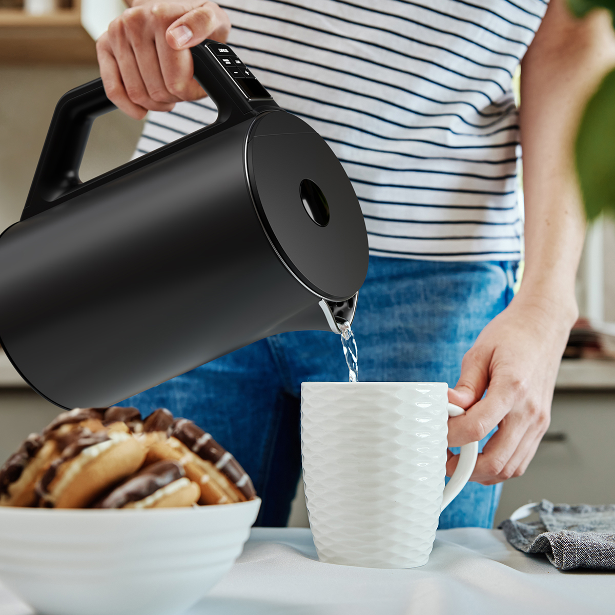 Eine Person gießt Wasser aus einem schwarzen Wasserkocher in eine weiße Tasse.