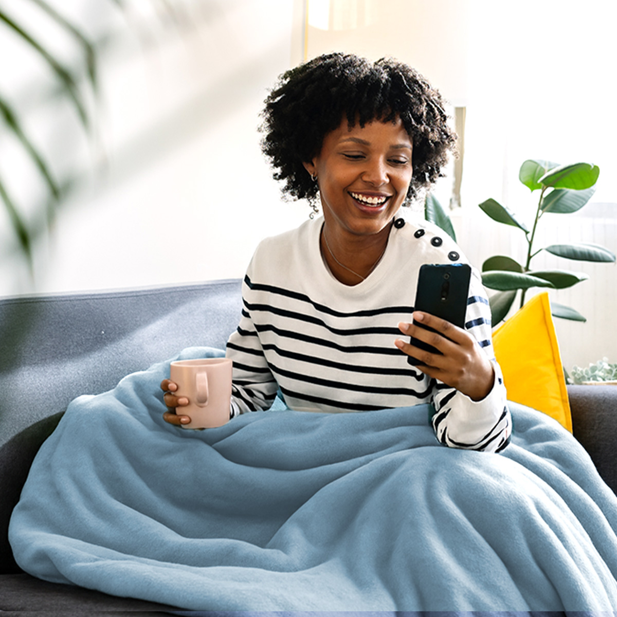 A woman with dark curly hair sits on a couch, wrapped in a blue blanket, holding a mug and phone.