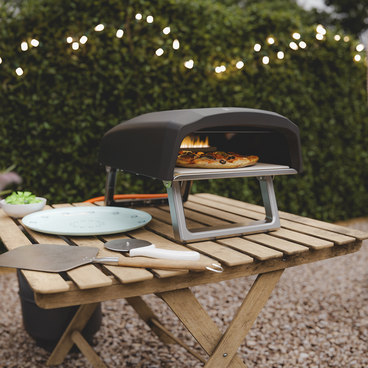 A pizza oven on an outdoor wooden table with a pizza inside and string lights in the background.