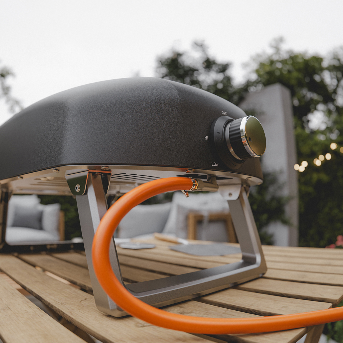 A black pizza oven with an orange hose sits on an outdoor wooden table.