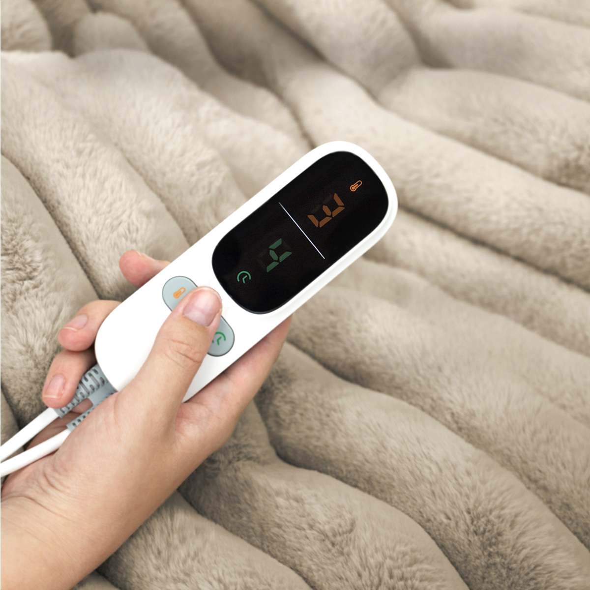 Hand holding a white electric blanket controller with a digital display on a beige fluffy blanket.