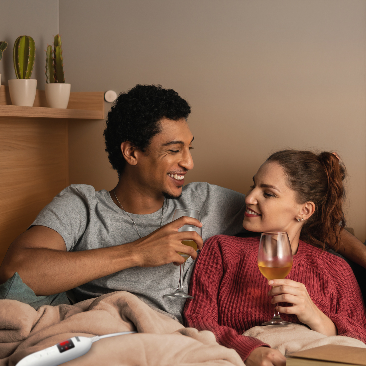 A couple smiles at each other, holding glasses of white wine, under a blanket.