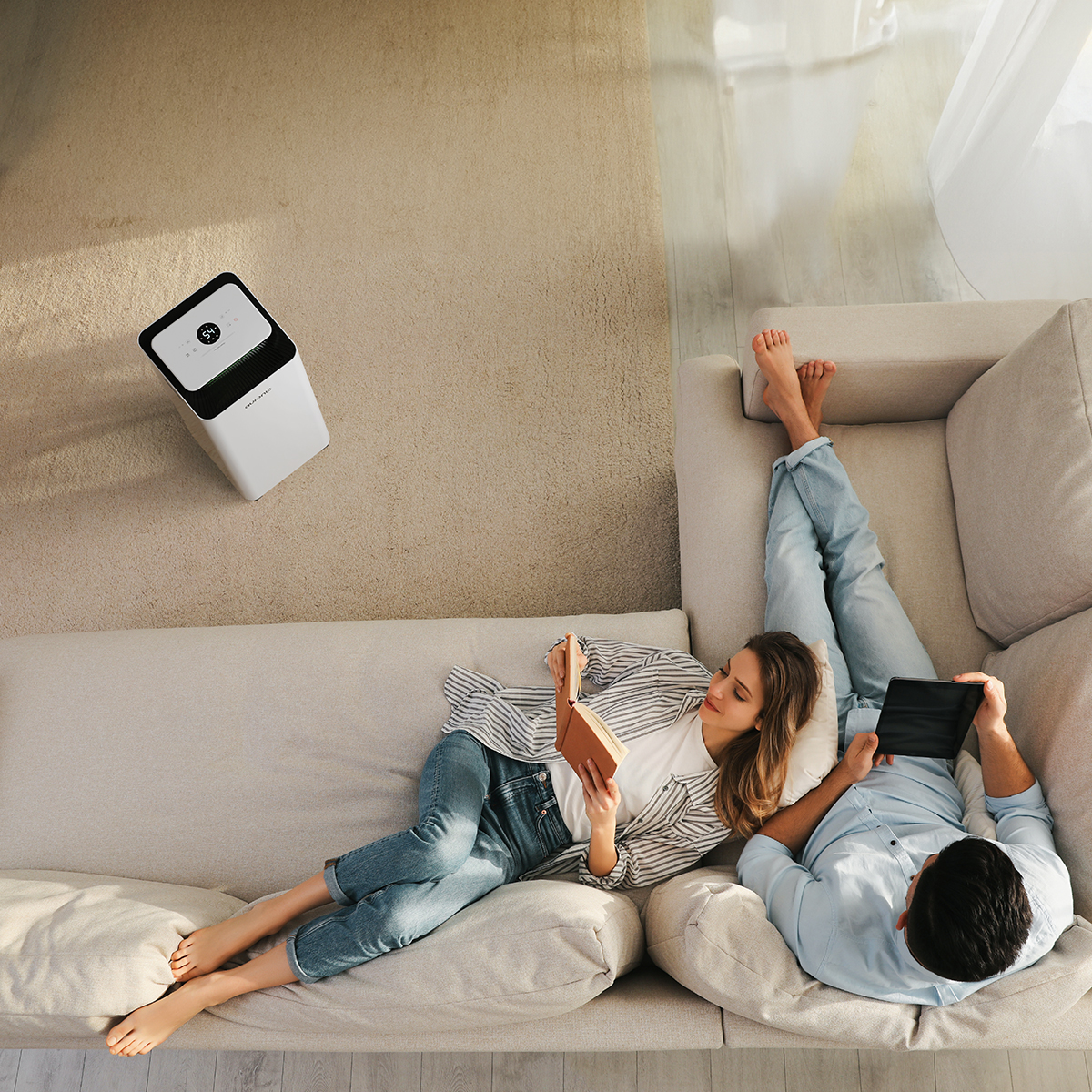 Un couple se détend sur le canapé, l'un lit un livre, l'autre une tablette. Purificateur d'air.