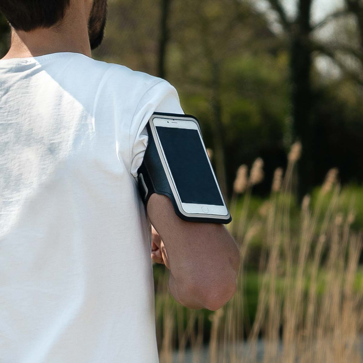 Een man met wit shirt draagt een telefoonarmband. Bomen en gras op de achtergrond.