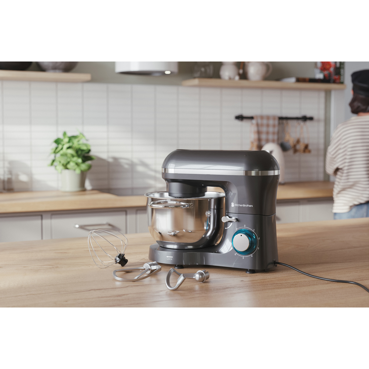 Robot pâtissier gris avec bol et accessoires sur table en bois, personne derrière.