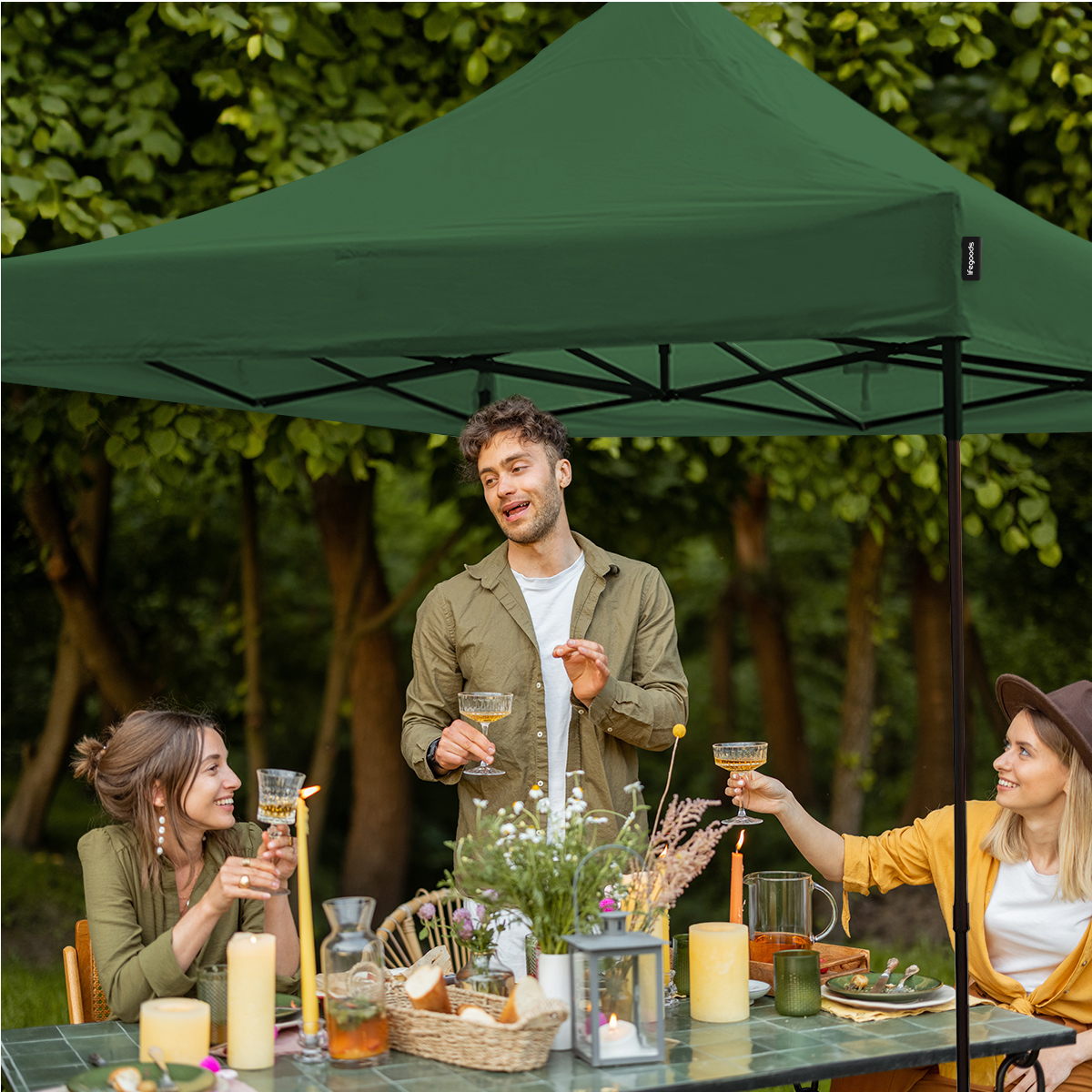 Three people celebrate under a green gazebo with glasses and food on the table.
