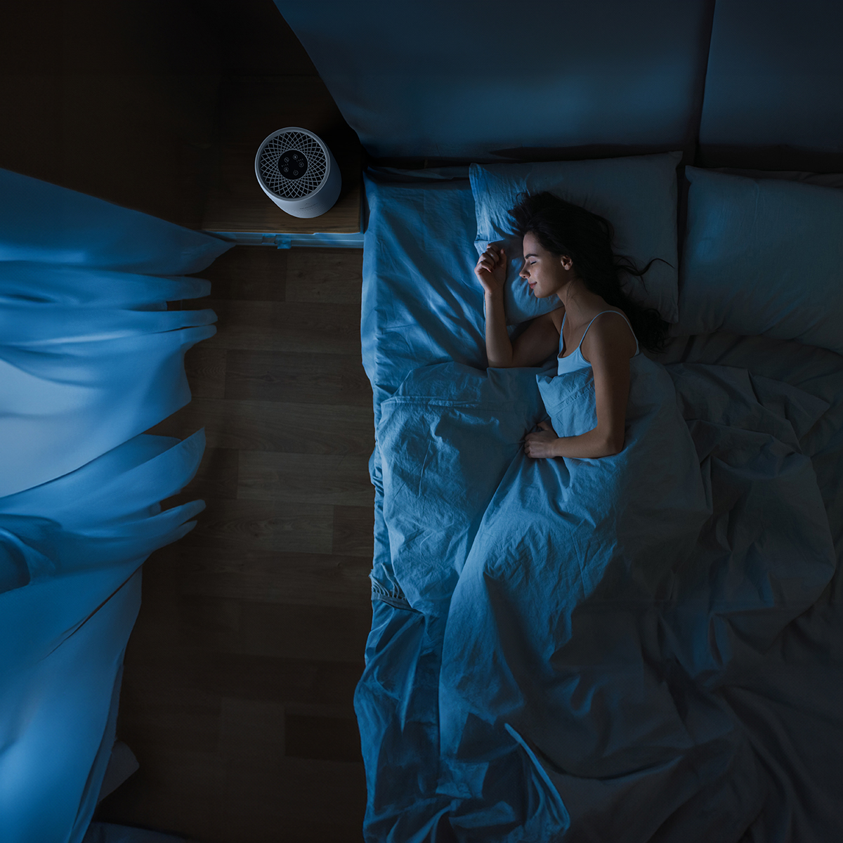 A woman sleeps in bed next to a nightstand with a fan.