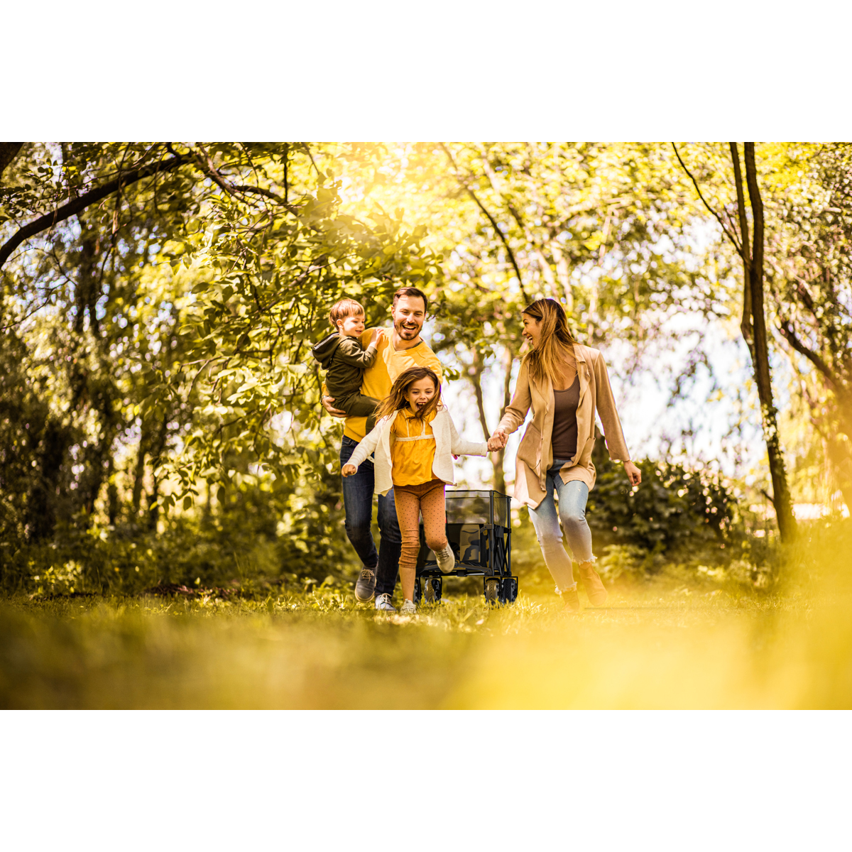 Eine Familie spaziert im Sonnenschein durch einen Park und zieht einen Wagen.