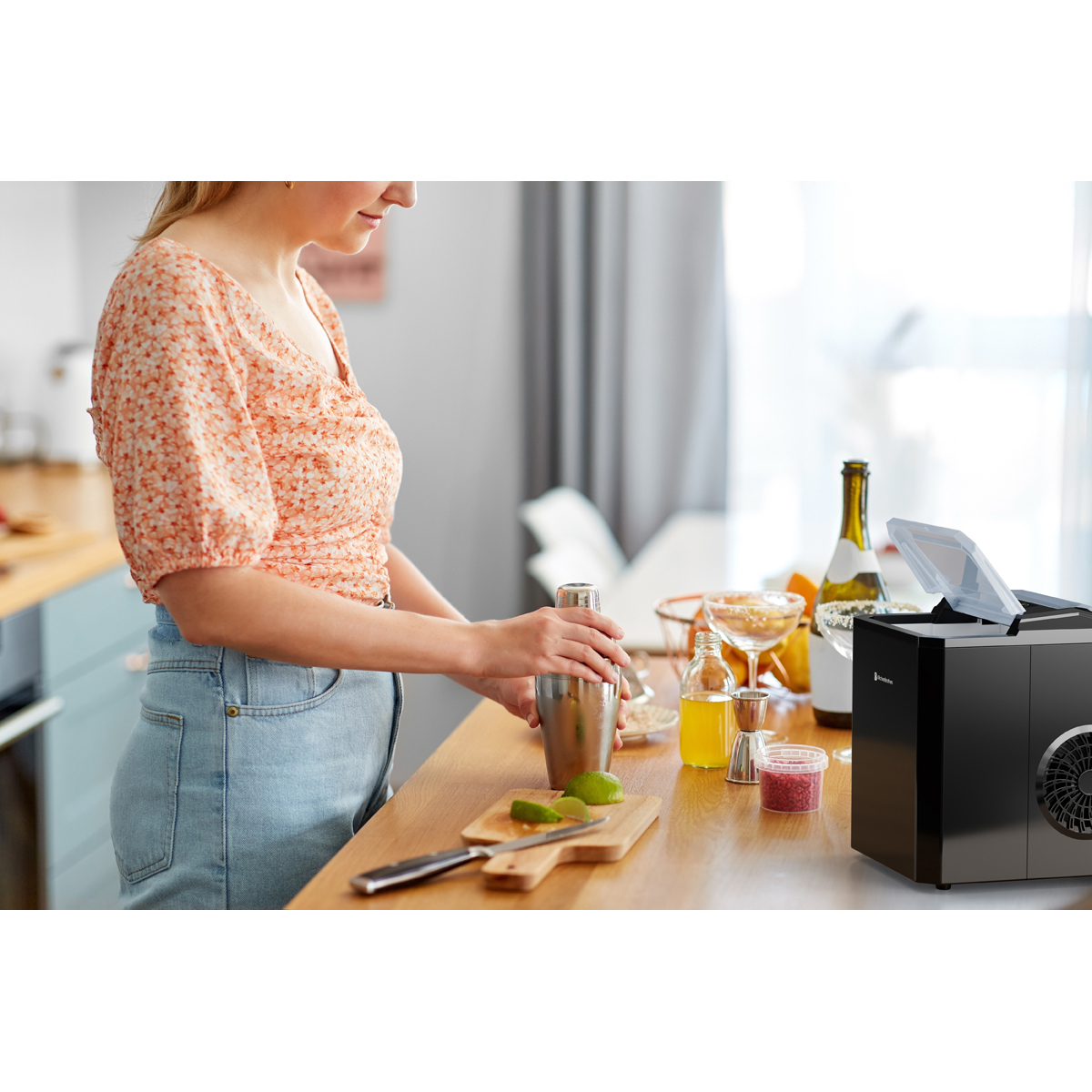 A woman mixes drinks with a shaker. Limes and an ice maker are on the table.