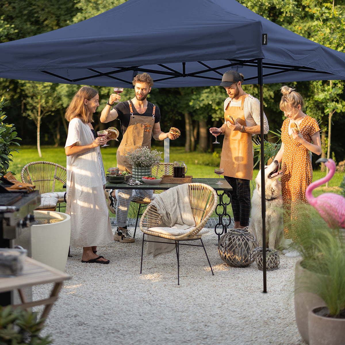 People enjoy BBQ under a navy canopy. A dog, chairs, table with food, and flamingo decor.