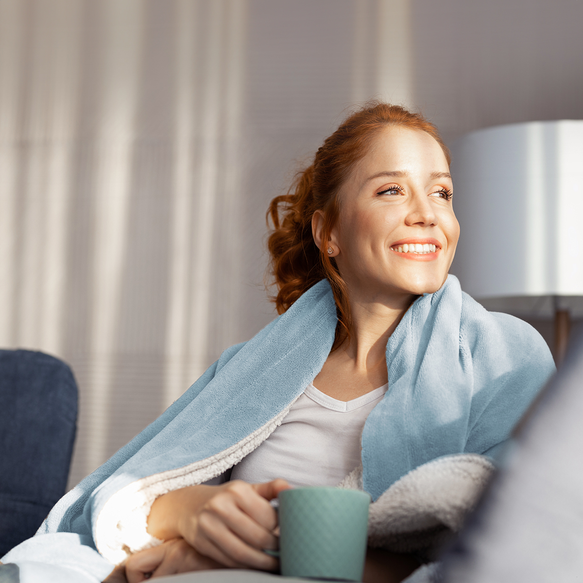 Une femme aux cheveux roux sourit, tenant une tasse, enveloppée dans une couverture bleue.