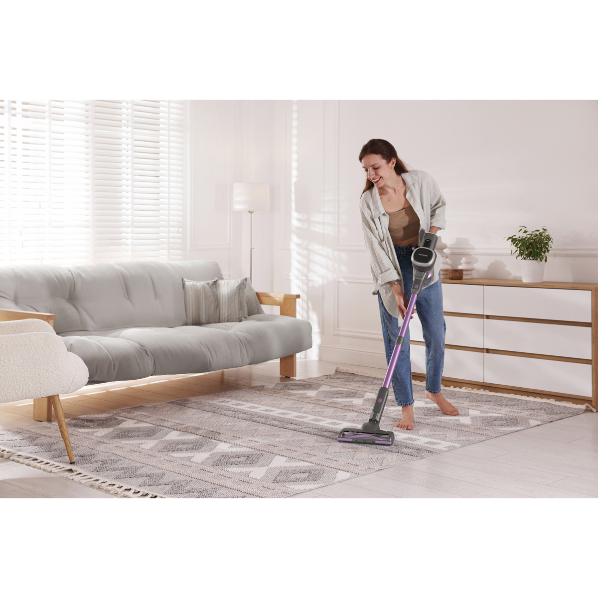 Une femme passe l'aspirateur sur un tapis avec un aspirateur balai violet dans un salon.