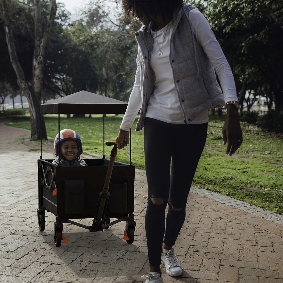 A woman pulls a wagon with a smiling child wearing a helmet on a park path.