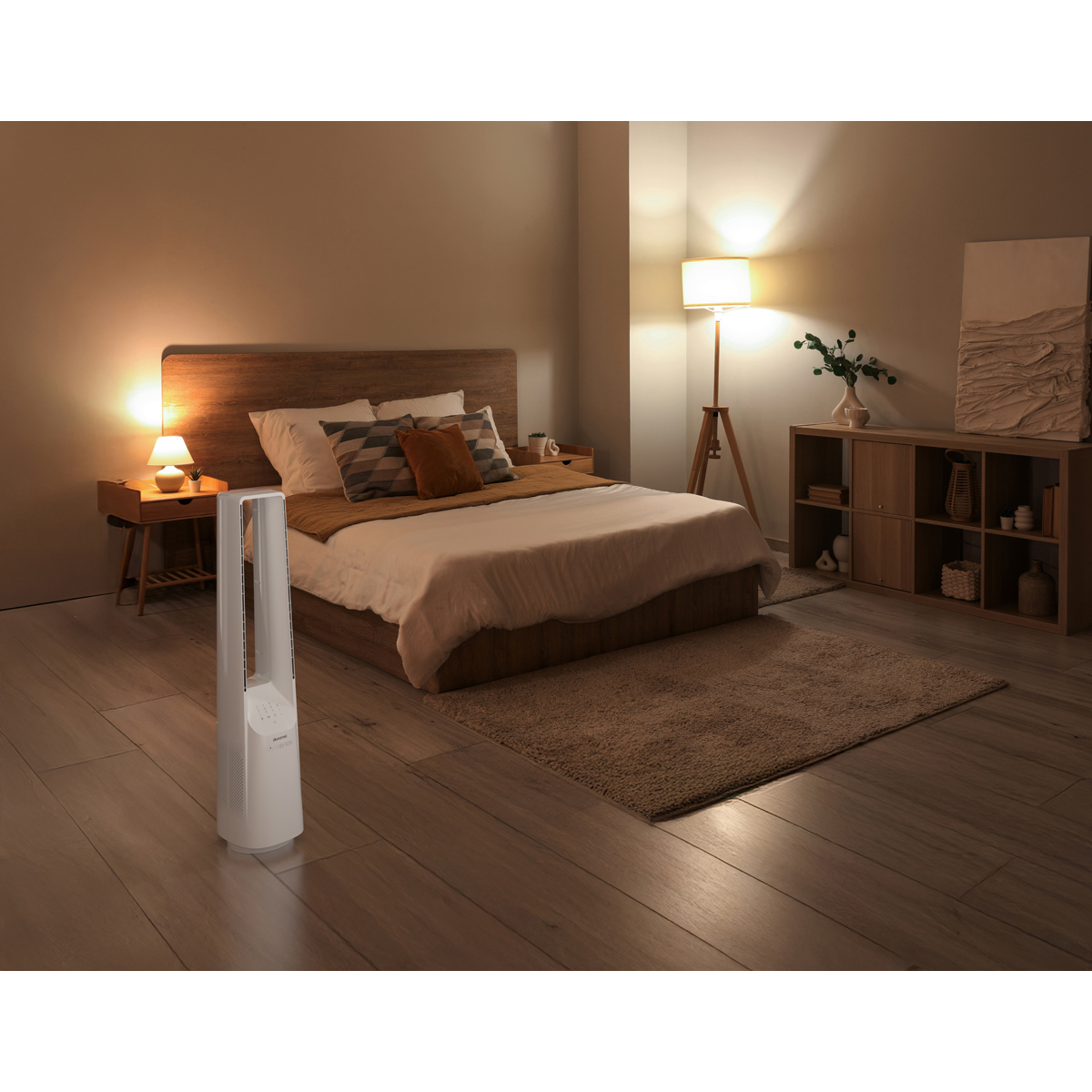 Ein weißer Turmlüfter steht auf einem Holzboden neben einem Bett in einem schwach beleuchteten Schlafzimmer.