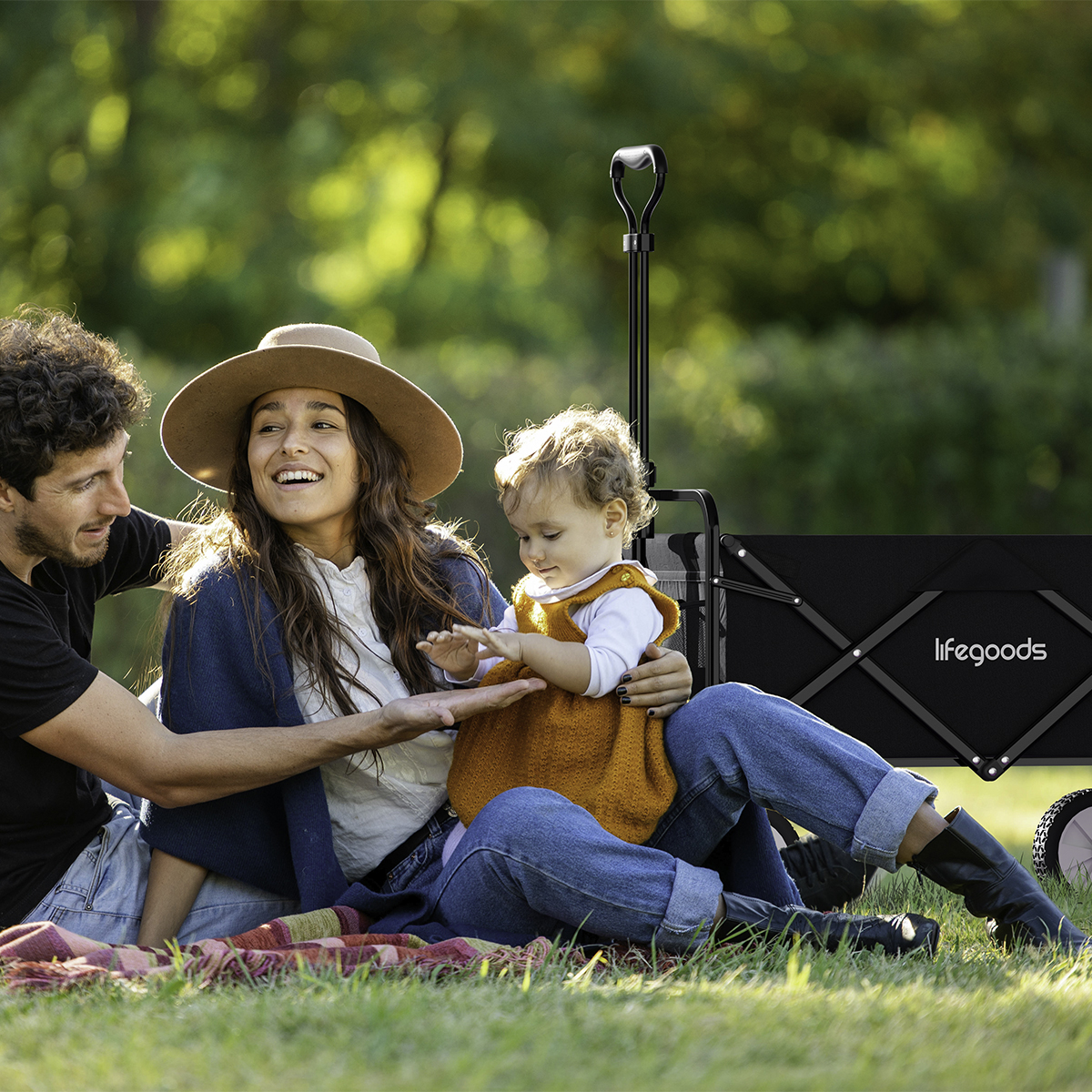 A family with a toddler sits on grass next to a black wagon.