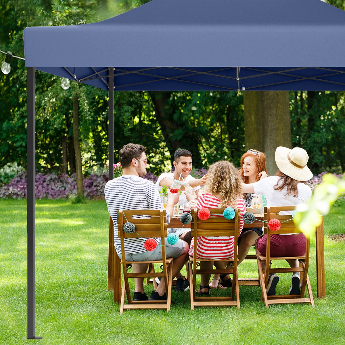 Friends enjoy a meal under a blue canopy in a sunny garden, seated at a wooden table.