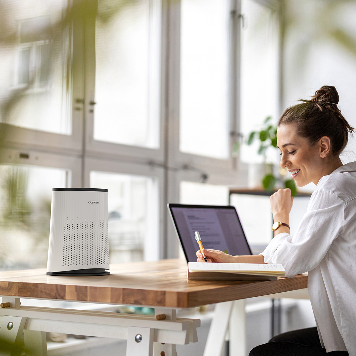 Woman at desk with laptop, air purifier, and notebook. Sunlight streams in.
