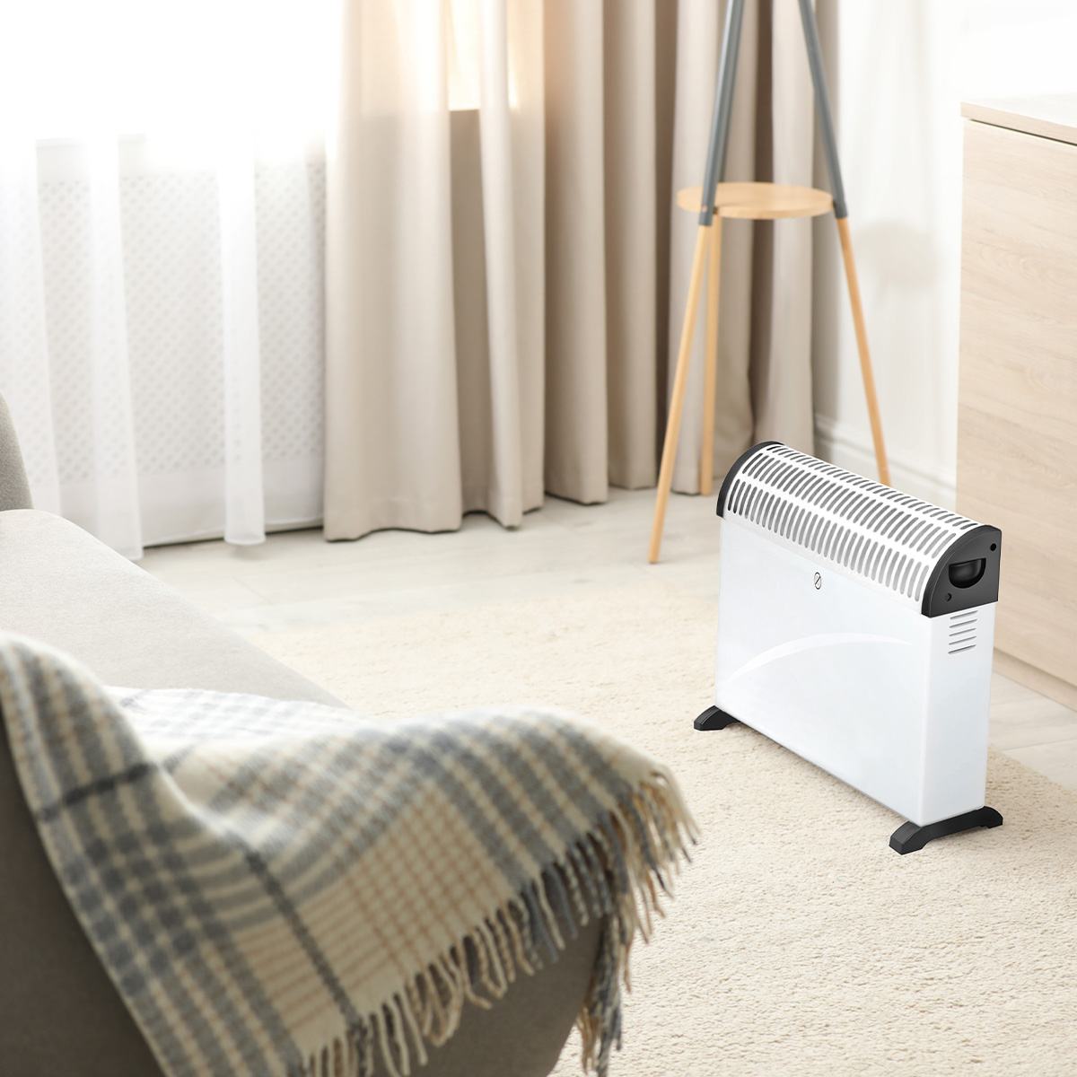 A white electric heater with black accents sits on a cream rug near a sofa with a plaid blanket.