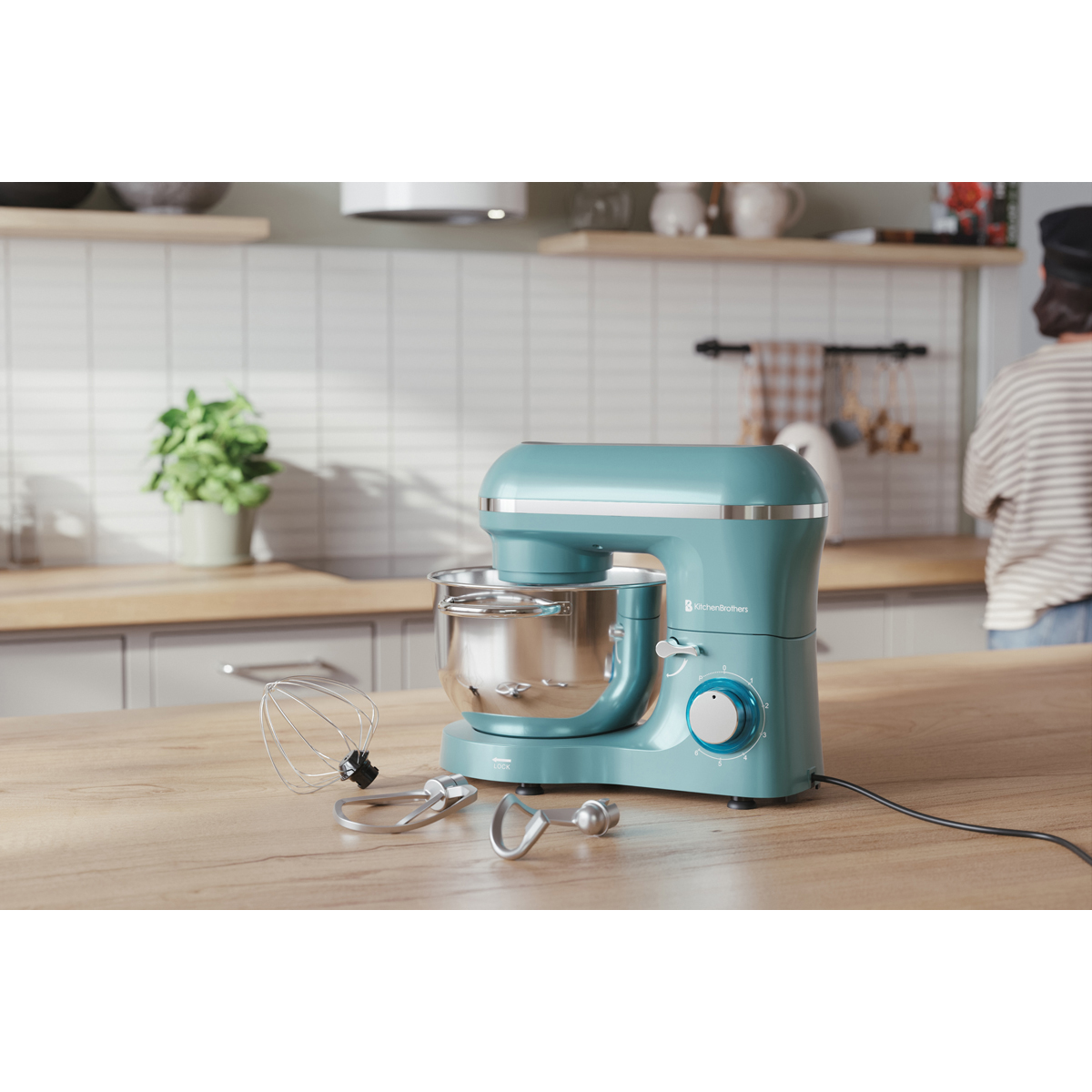 Een turquoise keukenmachine met accessoires op een houten aanrecht. Een persoon op achtergrond.