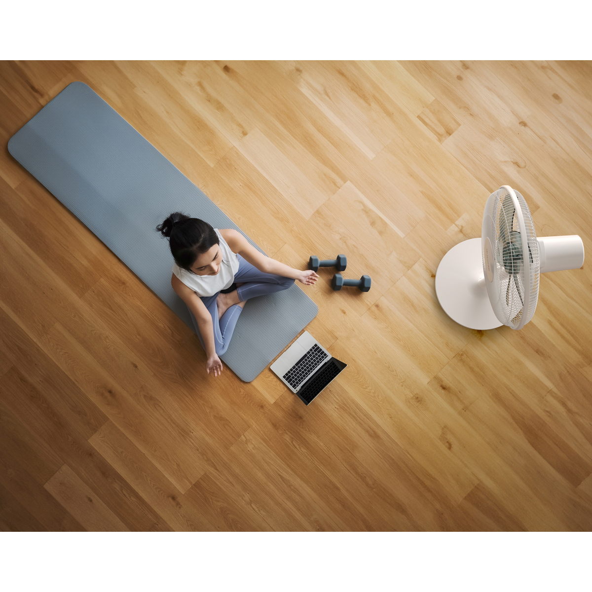 Femme assise sur un tapis, à côté d'haltères et d'un ordinateur portable. Ventilateur à droite.
