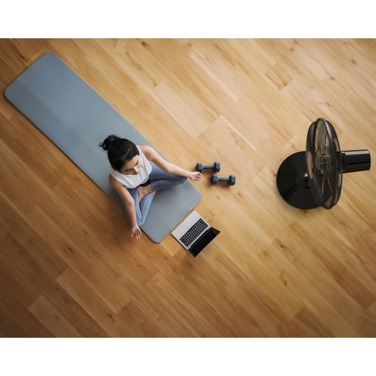 Femme méditant sur tapis de yoga avec haltères, ordinateur portable et ventilateur.