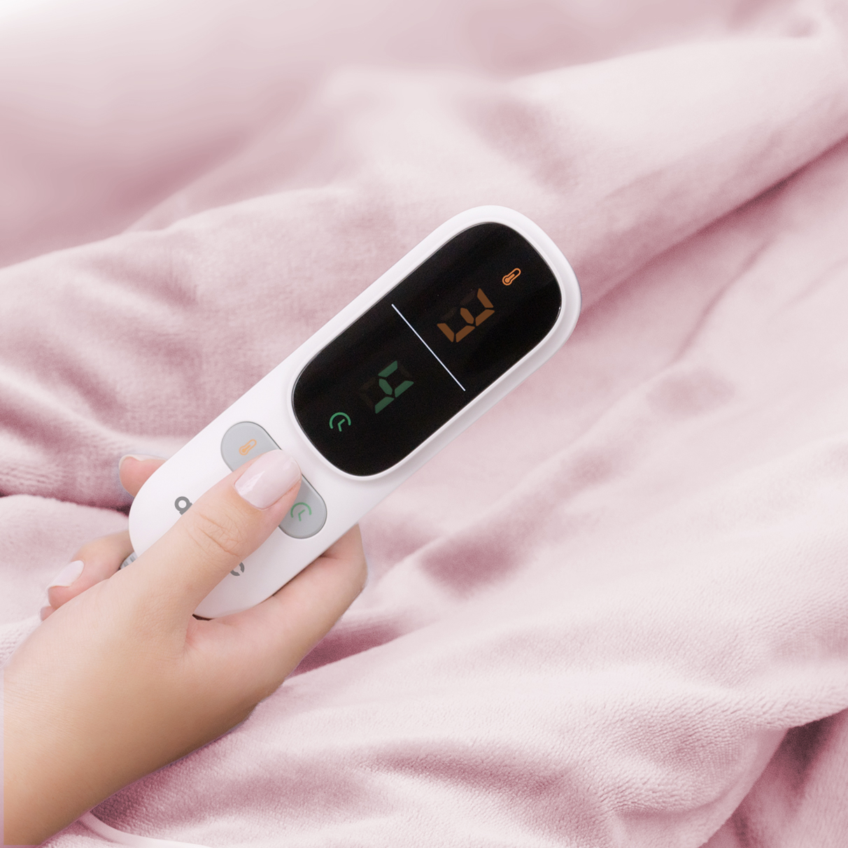 A hand holds a white digital thermometer against pink fabric. The display shows numbers.