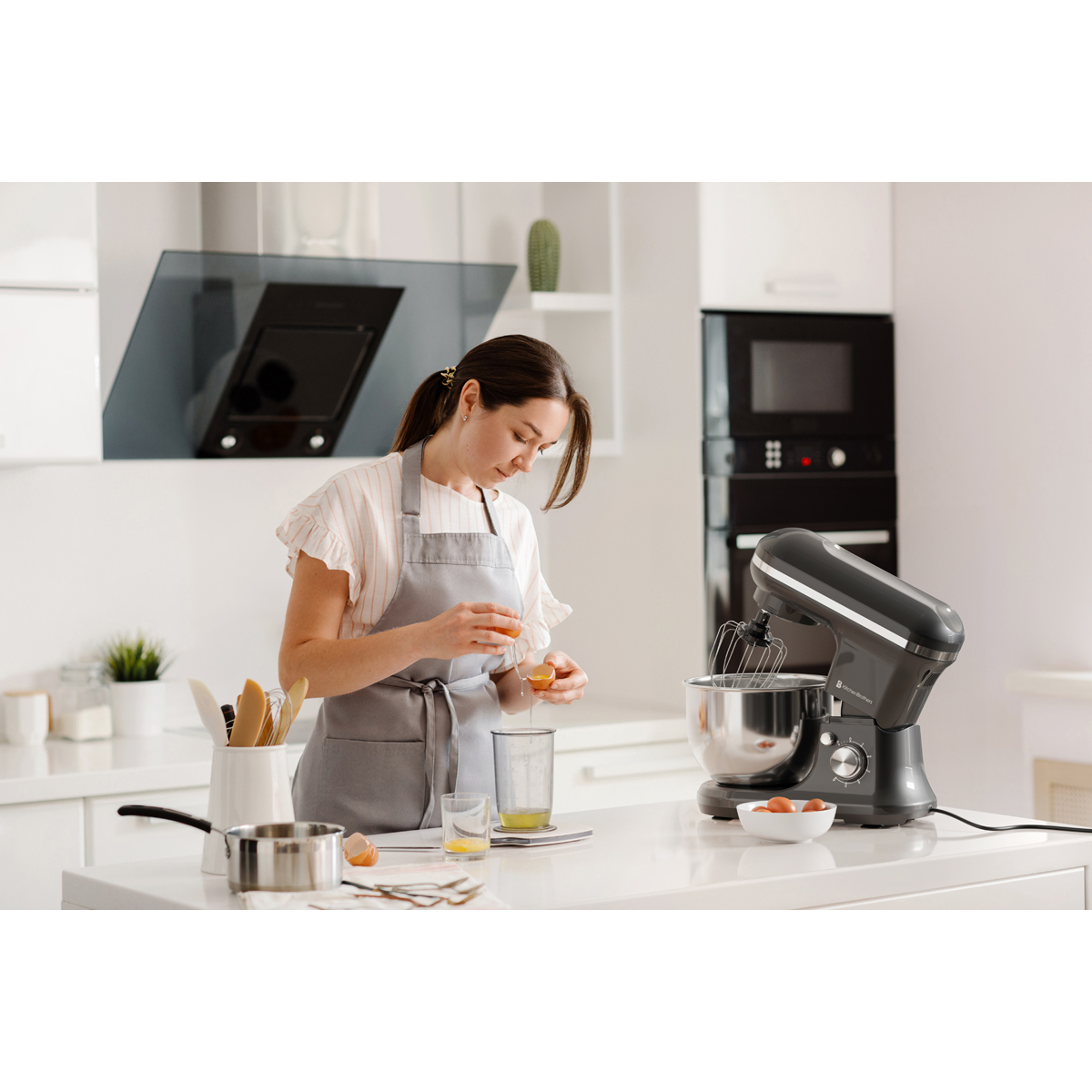 Une femme casse des œufs dans une cuisine moderne à côté d'un robot pâtissier.